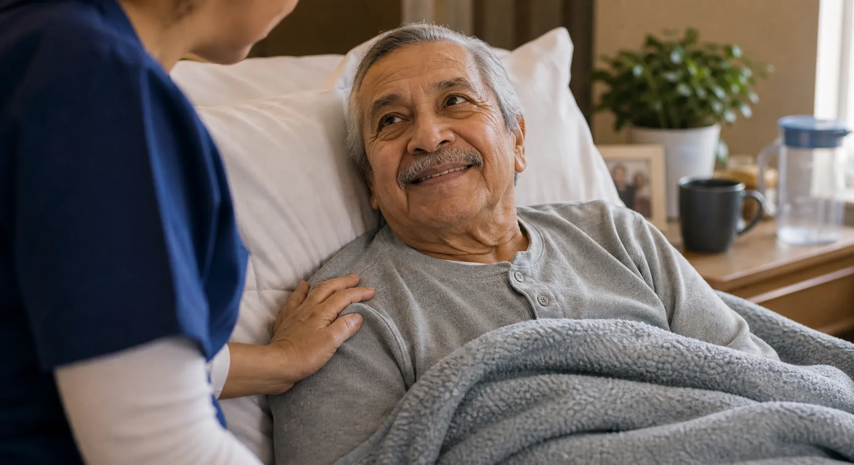 A nurse leans in to check on an elderly man resting comfortably in his bed